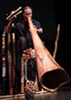 Dark background behind a man blowing a very large horn made of wood with pine cones around the horn.