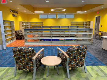 Two brown chairs with blue, peach, and yellow shapes are in the foreground of the photo with a small round table between them. The shelving for the children's area is in the background surrounded by bright yellow walls.
