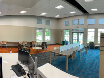 A panoramic view of the area with study tables, a shelving area for magazines, and the check out desk. The carpet is in sections with orange, then gray, and then blue. 