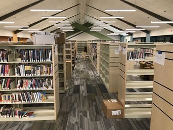 The area near the service desk in the adult area of the library. There are many shelving secionts with books, and some shelves that are still empty. There are lights hanging overhead from the ceiling. The carpet is gray, tan, and white.