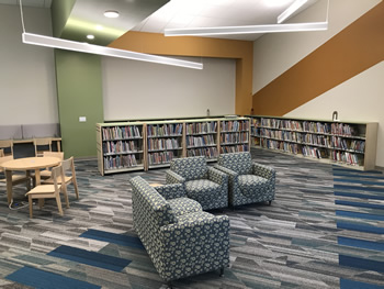 There are two child sized padded chairs and a child sized padded couch upholstered in a blue and white with circular designs. The carpet is blue, gray, tan, and white in a varigated pattern. There are books on bookshelves against the wall and free-standing. There is a round wooden table with wooden chairs to the left of the padded chairs. The walls are tan with one beam in green and another beam across the top and side in orange. There is a wide diagonal orange stripe on the wall to the right.