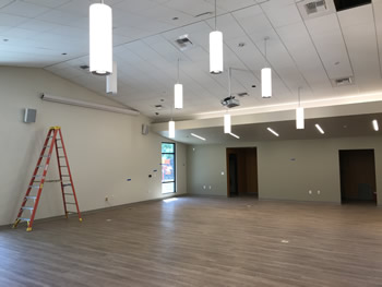 The Community Room with lights hanging down from ceiling and the wood flooring completed. The walls are tan and ceiling is white. There is a window in the back corner of the room.
