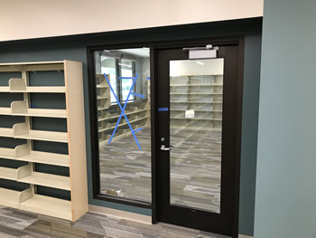 The window and glass entry door at the entrance to the Friends of the Turlock Public Library bookstore. There is shelving to the left of the entrance. The walls on the outside of the bookstore are green, with tan walls inside and shelving around the walls.