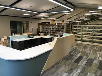 The service desk in the adult area of the library. The front of the desk is aqua colored with a white counter area. There is a light wood colored section around the side. There is shelving behind the desk. There are lights hanging overhead from the ceiling. The walls are tan. The carpet is gray, tan, and white.