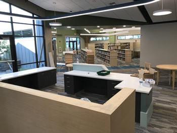 The view is from the back of the service desk in the children's area of the library. The back of the service desk area is a light wood colored section. The desk in front of that has black drawers and the side is aqua and white with a white counter area. There is shelving across the way in front of the desk. There are lights hanging overhead from the ceiling. The walls are tan, with green walls on the opposite side. The carpet is gray, tan, and white.