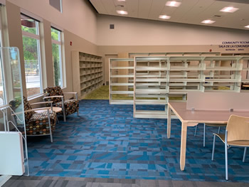Shelving for the books in the adult section, with a blue carpet, with gray triangles, two chairs, as well as a study table with chairs. There are two tall, narrow windows to the left behind the two chairs.