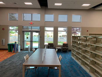 The inside view of the front entrance glass doors with windows to both sides of the doors, as well as small windows above the doors. The doors have RFID gates on each side. There is a set of shelving to the right, with a study table with chairs to the left of the shelving. The carpet is a section of orange to the left, gray in the middle section, and blue on the right section under the shelving.