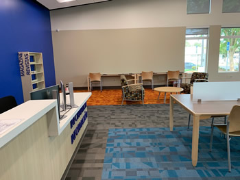 A panoramic view of the area with study tables, a shelving area for magazines, and the check out desk. The carpet is in sections with orange, then gray, and then blue. 