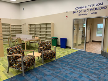 Two brown chairs with blue, peach, and yellow shapes are in the foreground of the photo with a small round table between them. The shelving for the teen area, as well as a study area table, is in the background. The walls are beige.