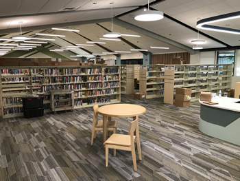 The area near the service desk in the adult area of the library. There is a round wooden table with 2 chairs near the shelving area. There are several shelves with books, and some shelves that are still empty. There are lights hanging overhead from the ceiling. The carpet is gray, tan, and white.