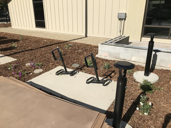 The side area of the main entrance to the Turlock Library with a cement area and two individual bike racks. The cement area is surrounded with bark mulch and some small plants. The tan wall of the library is seen in the background.
