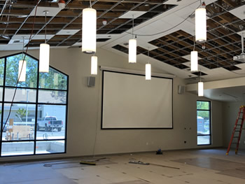The Community Room with lights installed from the ceiling hanging down, the projection screen on the wall with a large window to the left, and flooring in progress.