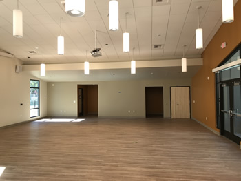 The Community Room with lights hanging from the ceiling, a window to the left, doors to the back, and light colored wood flooring. The glass double door entrance is to the right in an orange wall.